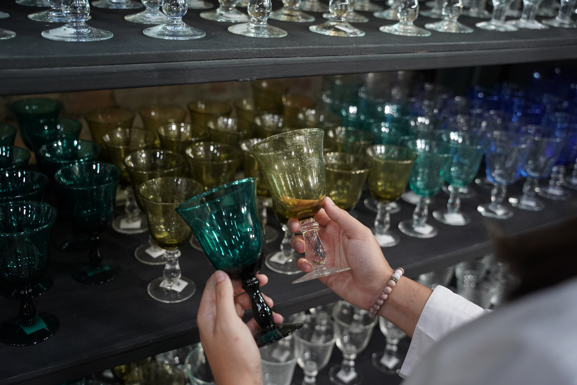 Hands holding teal and olive handblown goblets in front of a colour-graded shelf